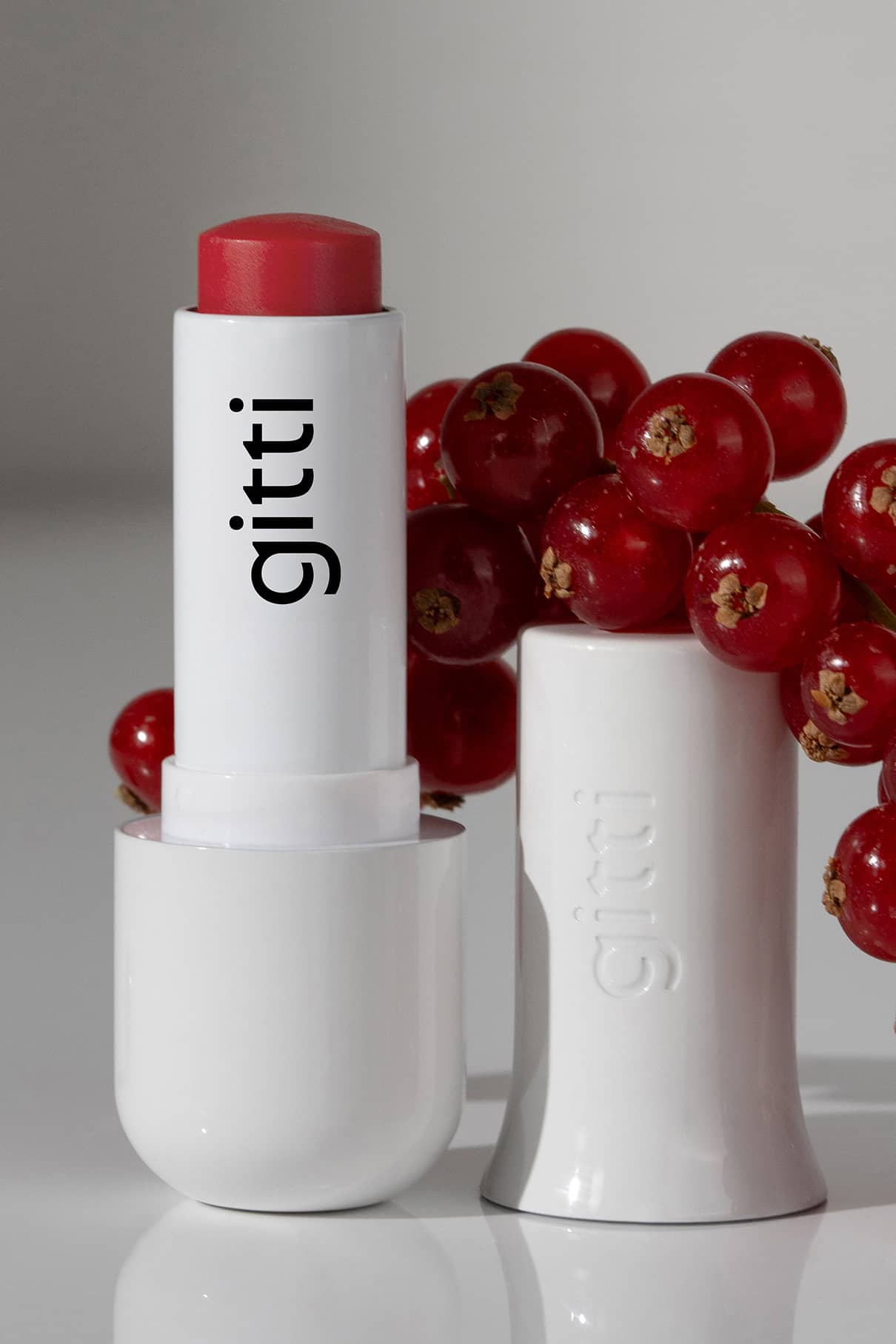 Ein geöffnetes Lip Sync Refill no. 02 steht neben seiner Kappe und roten Johannisbeeren auf einer spiegelnden Oberfläche - ideal für kühle Hauttöne mit seinem beerenroten Farbton und rosa Untertönen.