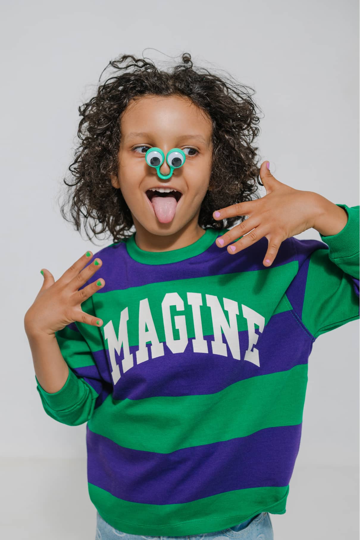 Ein Kind in einem grünen und lila IMAGINE-Sweatshirt posiert spielerisch mit Kulleraugen auf der Nase und herausgestreckter Zunge und zeigt die leuchtenden Farben des gitti kids no. 04 Nagellack.