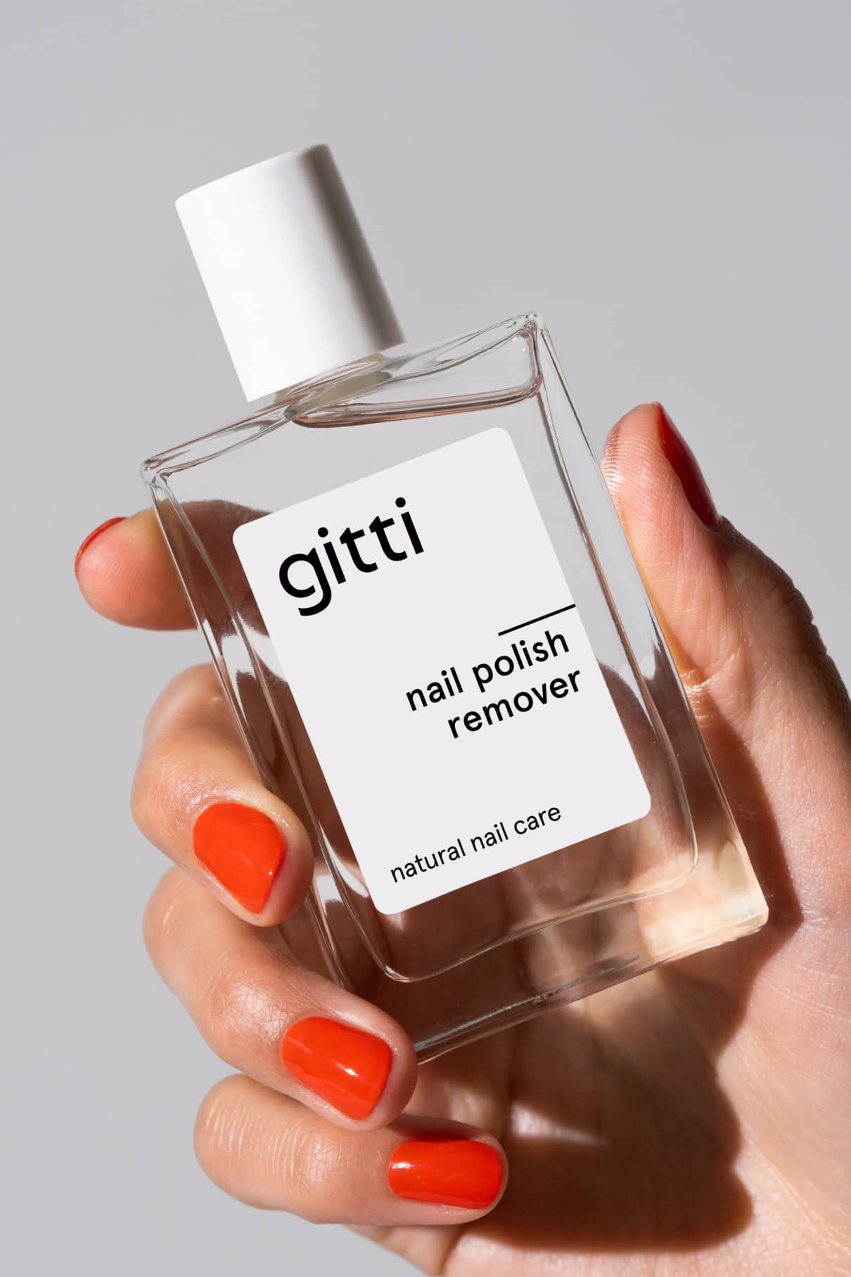 Eine orange-rot manikürte Hand hält eine Klarglasflasche mit der Aufschrift Nagellackentferner, ein natürlicher Nagellackentferner, der für seinen angenehmen Duft und seine sanfte Nagelpflege bekannt ist.
