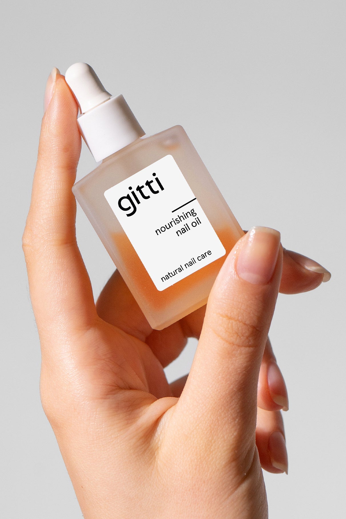 Eine Hand hält eine Flasche Be A Softie Nourishing Nail Oil mit einem Tropfverschluss mit der Aufschrift Natural Nail Care neben dem dazugehörigen Set vor einem schlichten hellgrauen Hintergrund.