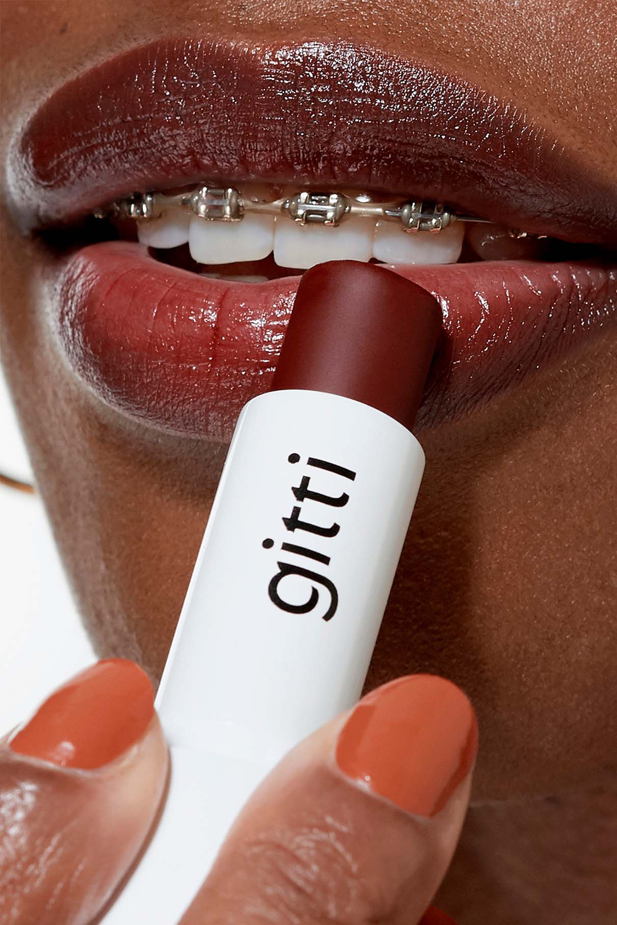 Eine Person mit Zahnspange trägt rötlich-braunen Lippenstift auf ihre Lippen auf; ihre orange lackierten Fingernägel sind Teil der Community Box, einem exklusiven Set mit kostenlosem Versand.