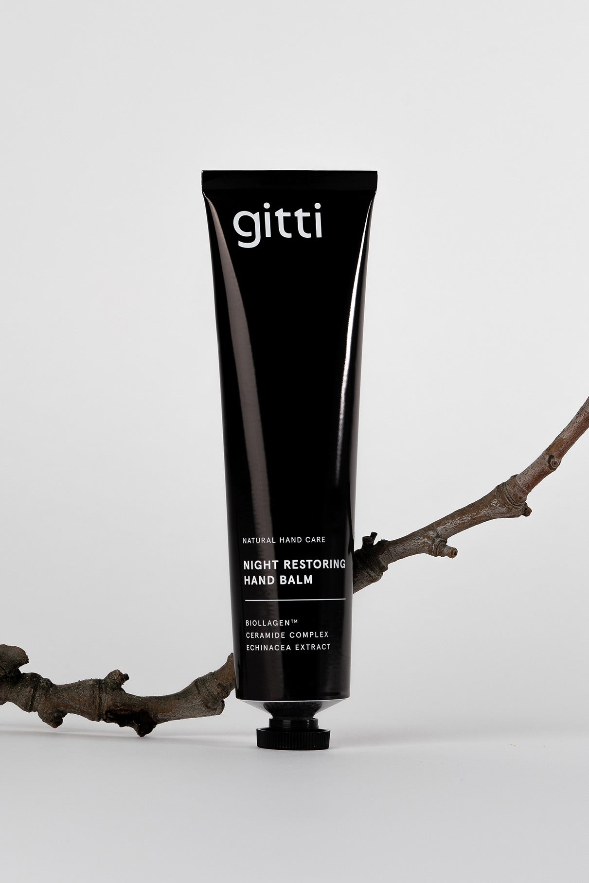 Eine schwarze Tube mit der Aufschrift gitti Be A Softie Hand Balm steht aufrecht neben einem Nourishing Nail Oil, beide ruhen auf einem kahlen Baumast vor einem schlichten weißen Hintergrund.